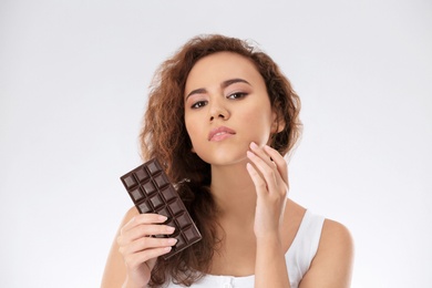 Beautiful young woman with acne problem holding chocolate on light background. Skin allergy