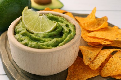 Bowl of delicious guacamole, lime and nachos chips on wooden board, closeup