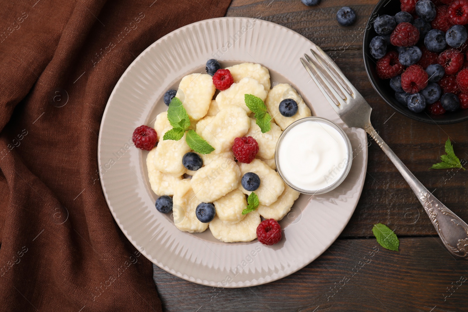 Photo of Plate of tasty lazy dumplings with berries, sour cream and mint leaves on wooden table, flat lay