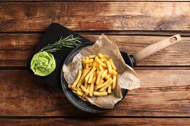 Serving pan with parchment, french fries, avocado dip and rosemary on wooden table, top view