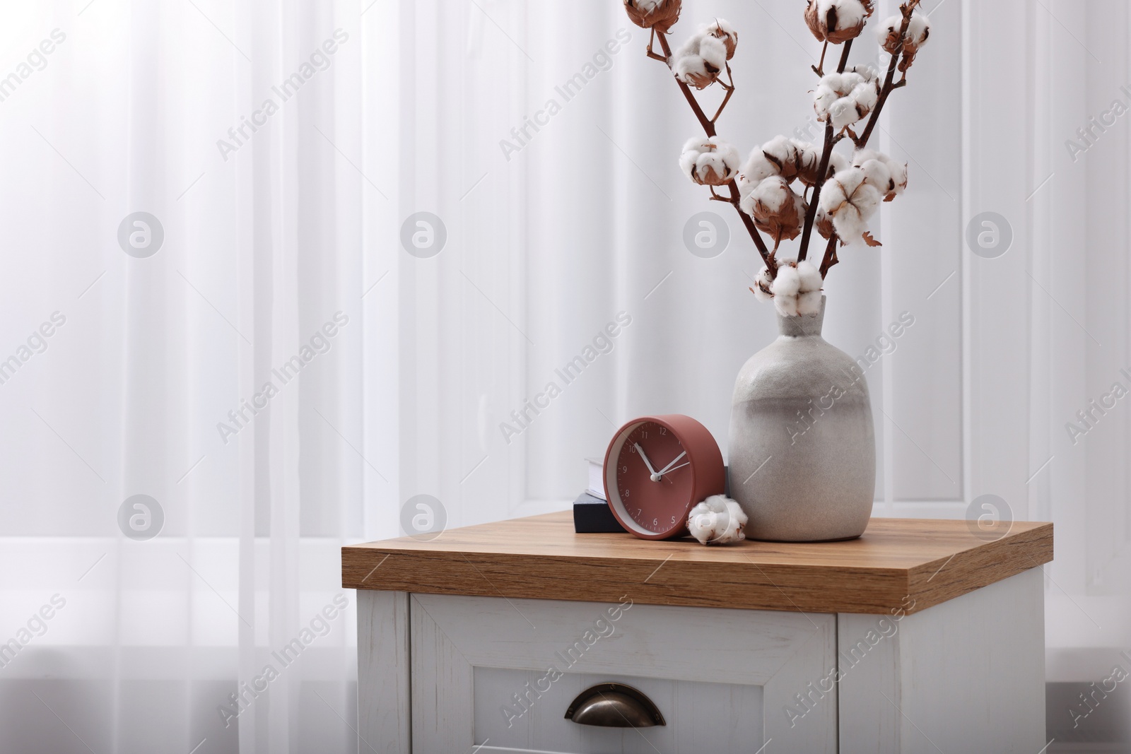 Photo of Alarm clock and cotton branches with fluffy flowers in vase on wooden commode indoors. Space for text