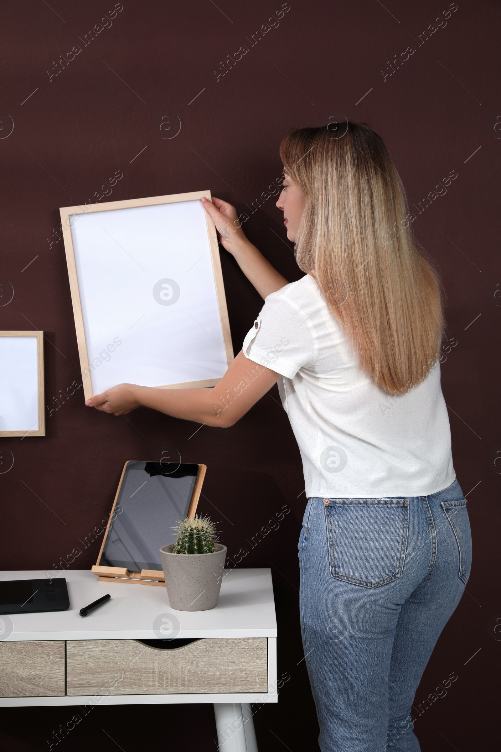 Photo of Young woman hanging empty frame on brown wall indoors. Mockup for design