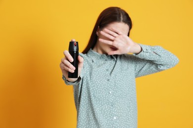 Young woman covering eyes with hand and using pepper spray on yellow background