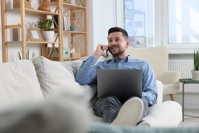 Photo of Happy man talking on phone while working with laptop on sofa at home