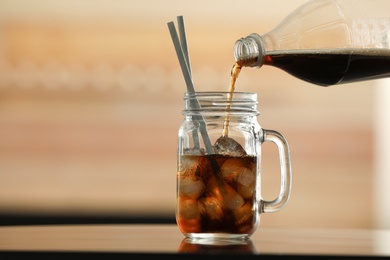 Photo of Pouring cola from bottle into mason jar with ice cubes on table against blurred background. Space for text