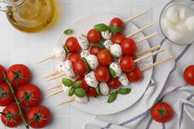 Photo of Caprese skewers with tomatoes, mozzarella balls, basil and spices on white tiled table, flat lay