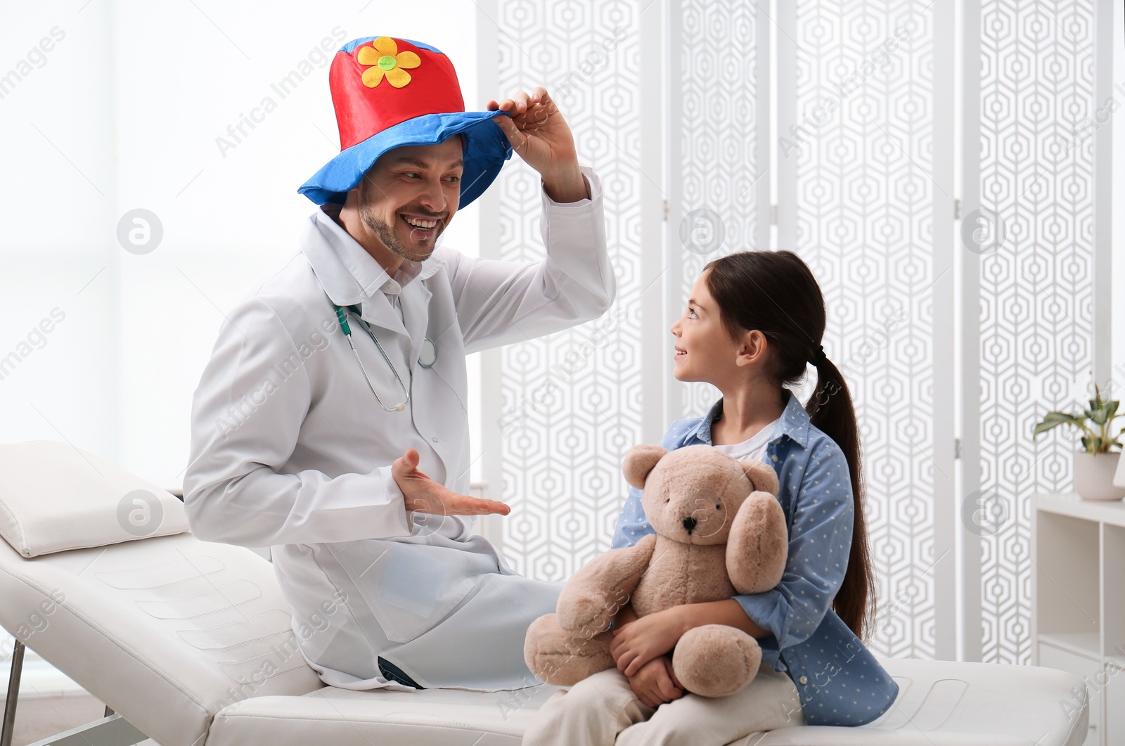 Photo of Pediatrician in funny hat playing with little girl during visit at hospital