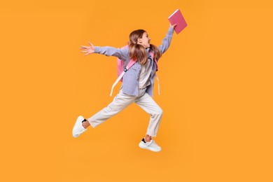Happy schoolgirl with book and backpack jumping on orange background