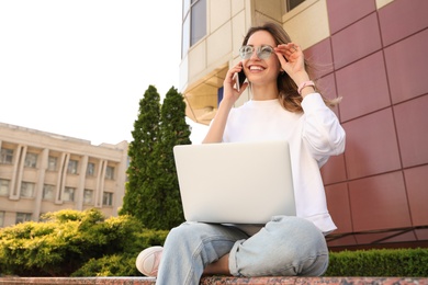 Happy young woman with laptop talking on phone outdoors