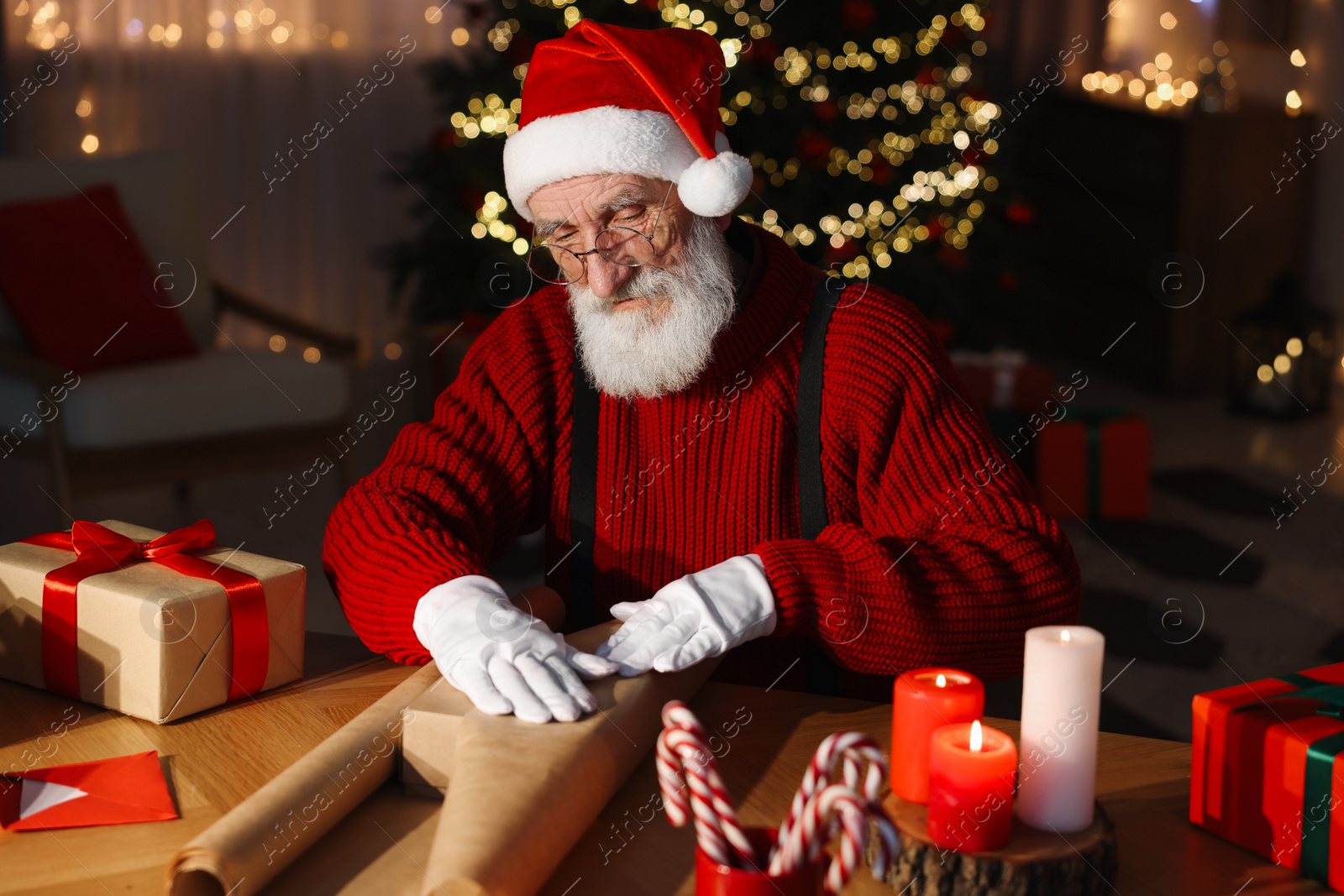 Photo of Santa Claus wrapping gift at his workplace in room decorated for Christmas