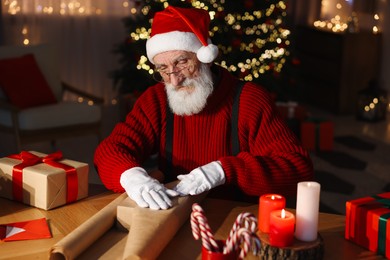 Santa Claus wrapping gift at his workplace in room decorated for Christmas