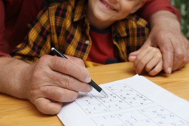 Photo of Little boy with his grandfather solving sudoku puzzle at wooden table, closeup