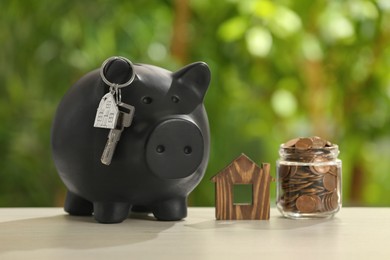 Piggy bank with key, house model and coins in glass jar on wooden table outdoors. Saving money concept