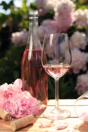 Photo of Bottle and glass of rose wine near beautiful peonies on wooden table in garden