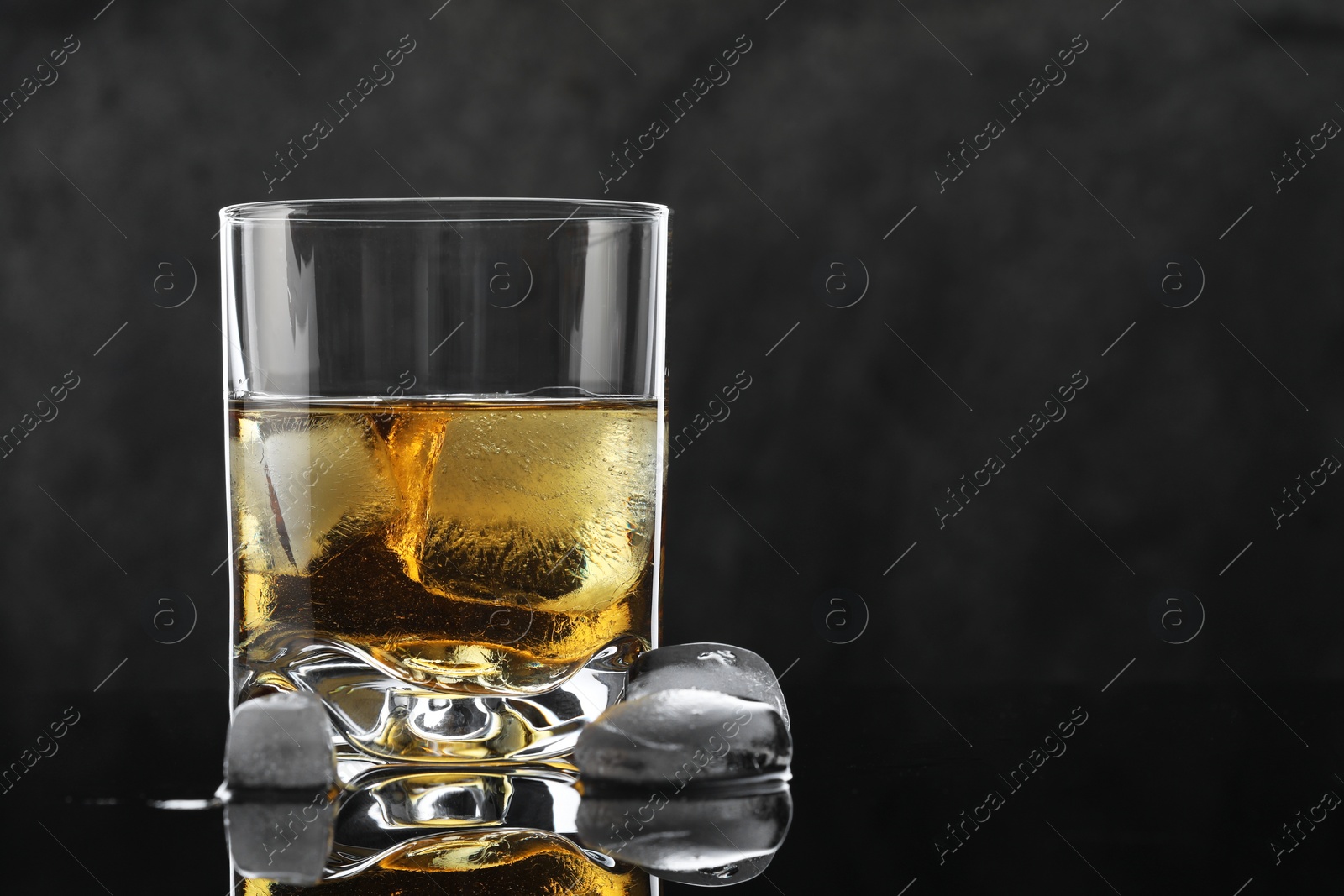 Photo of Tasty whiskey and ice cubes in glass on mirror table against black background, closeup. Space for text