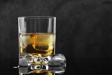 Tasty whiskey and ice cubes in glass on mirror table against black background, closeup. Space for text