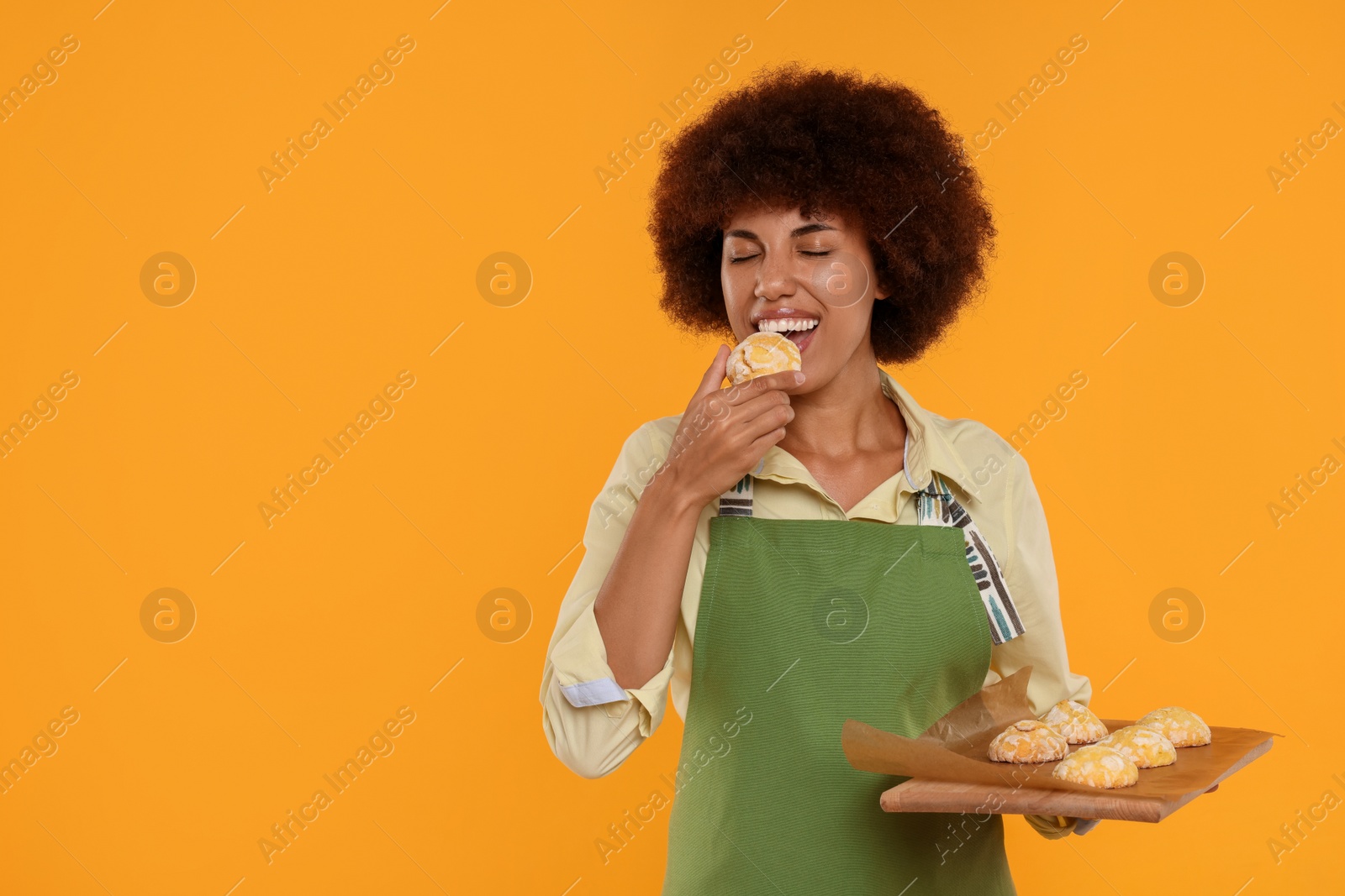 Photo of Happy young woman in apron holding board with cookies on orange background. Space for text