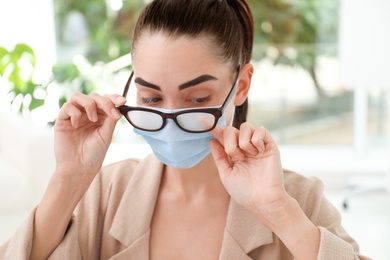 Woman wiping foggy glasses caused by wearing medical mask indoors, closeup