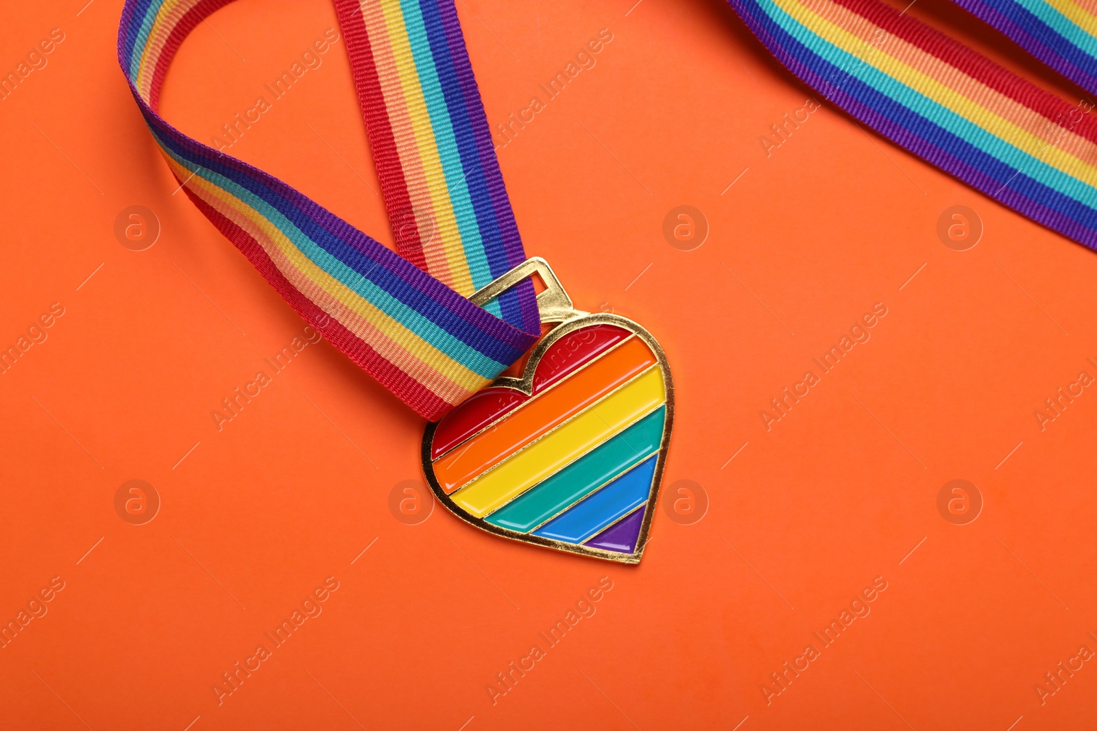 Photo of Rainbow ribbon with heart shaped pendant on orange background, top view. LGBT pride