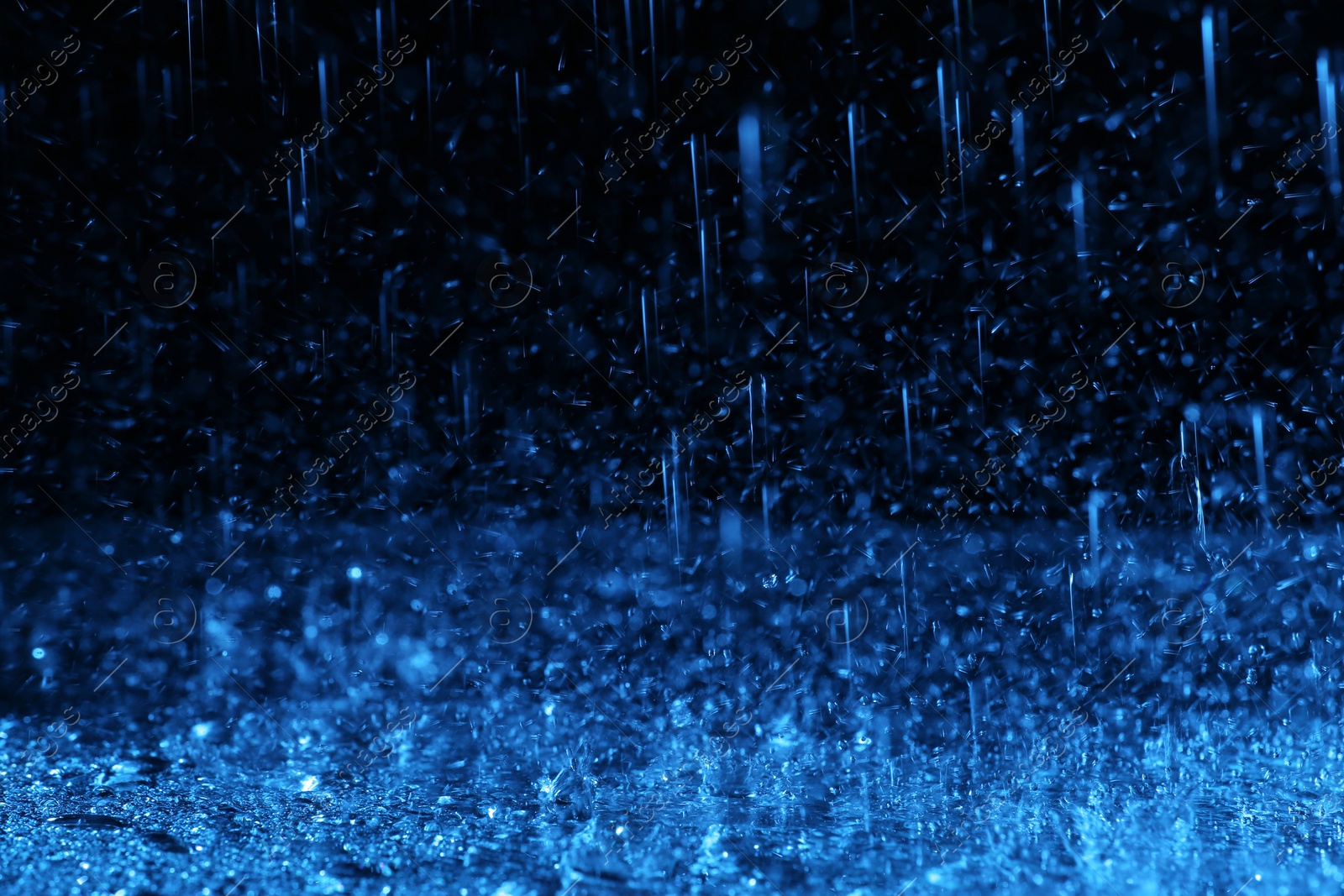 Photo of Heavy rain falling down on ground against dark background, toned in blue