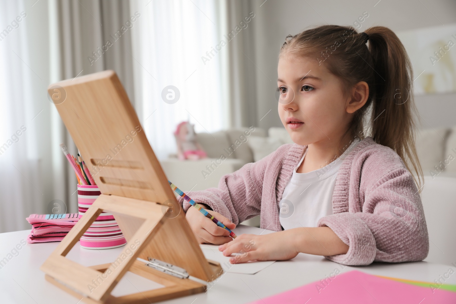 Photo of Adorable little girl doing homework with tablet at table indoors