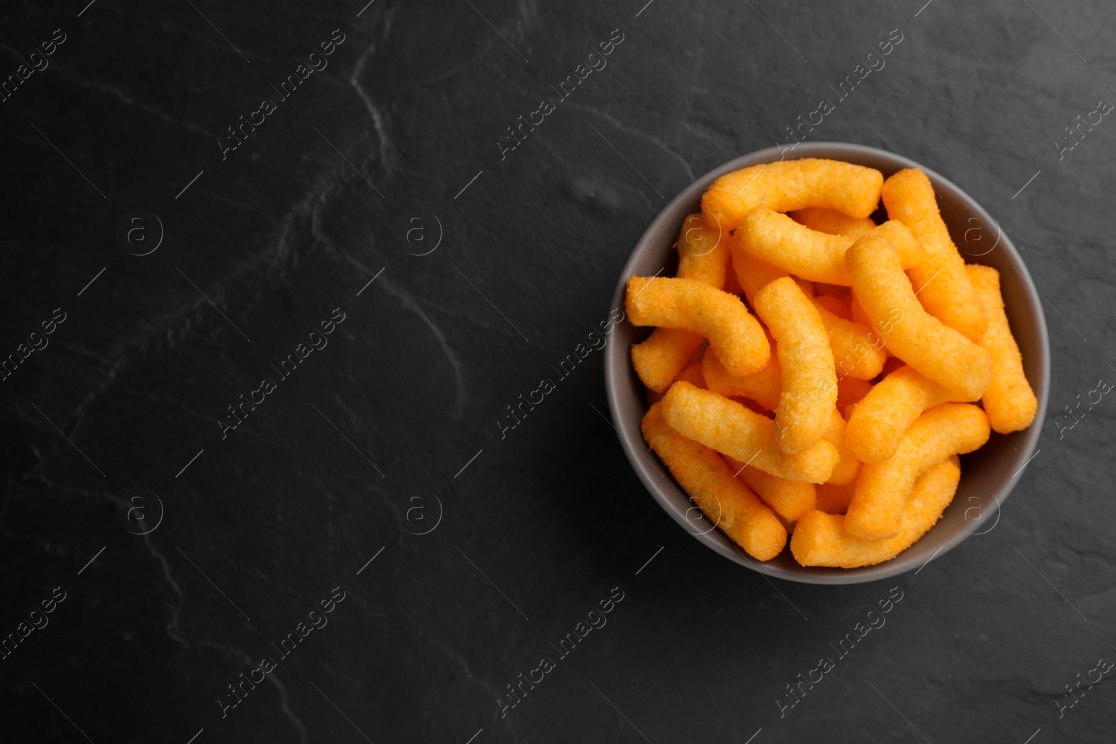 Photo of Many tasty cheesy corn puffs in bowl on black table, top view. Space for text