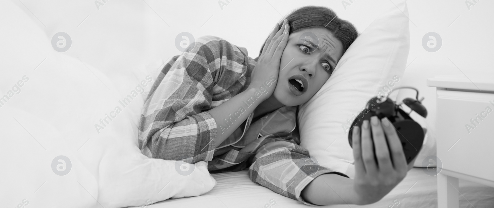Image of Emotional young woman with alarm clock at home in morning. Black and white photography