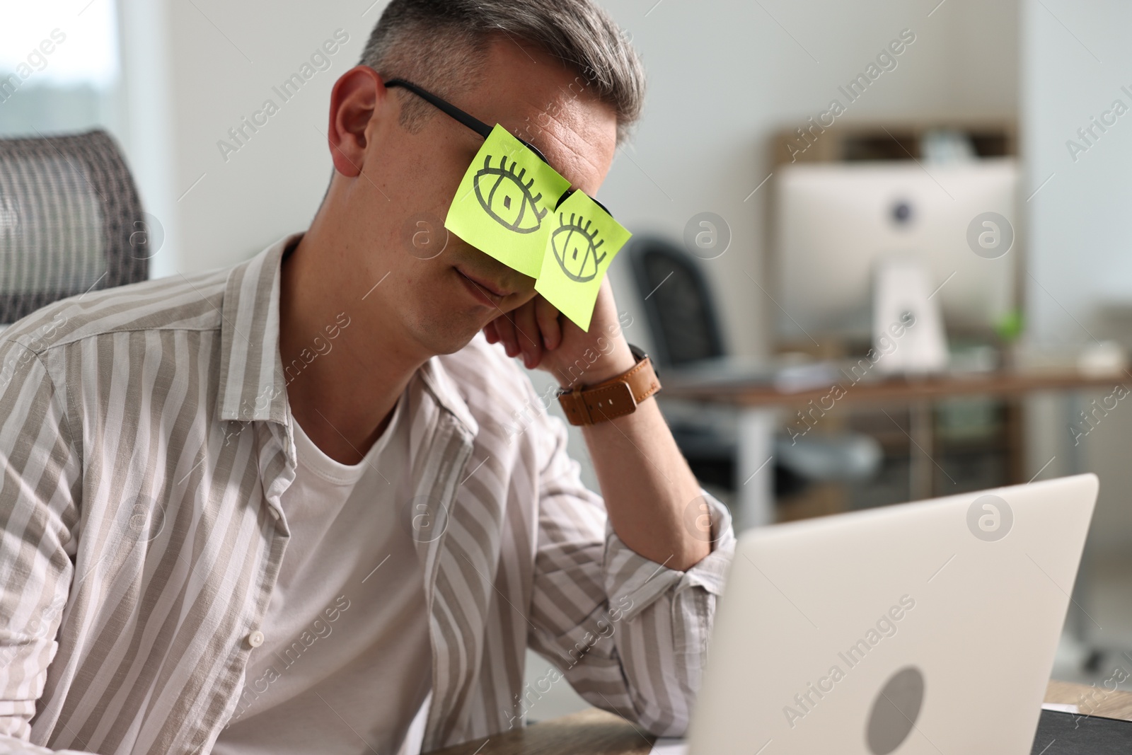 Photo of Man with fake eyes painted on sticky notes snoozing at workplace in office