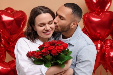 Photo of Happy couple celebrating Valentine's day. Beloved woman with bouquetred roses near heart shaped air balloons on beige background