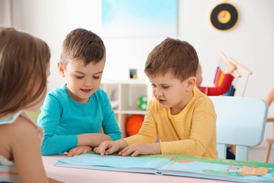 Photo of Cute little children reading book together at table indoors. Learning and playing
