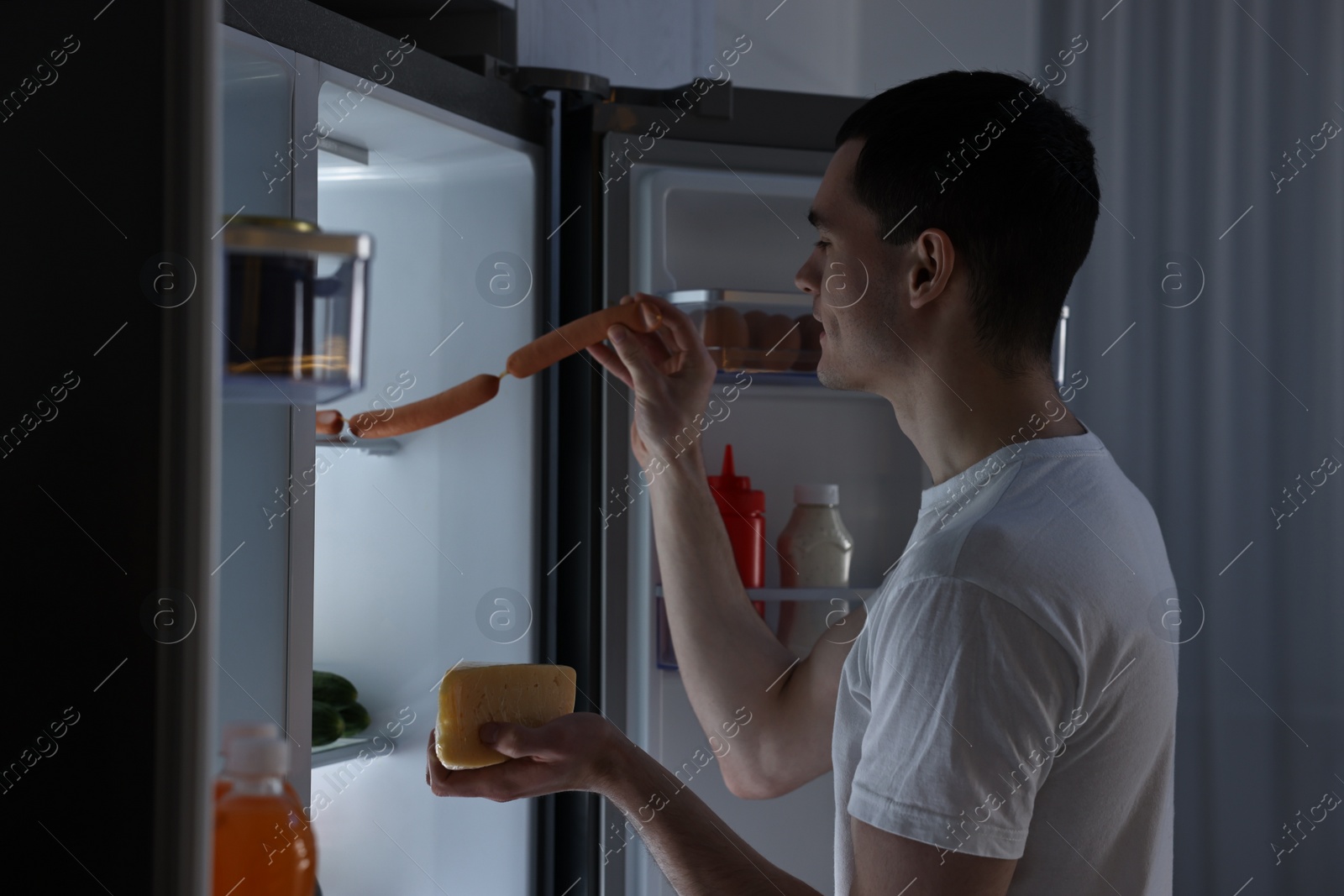 Photo of Man with cheese taking sausages out of refrigerator in kitchen at night