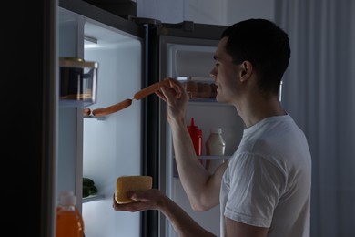 Man with cheese taking sausages out of refrigerator in kitchen at night