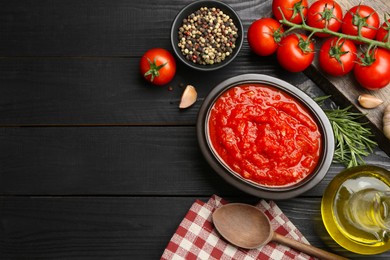 Photo of Homemade tomato sauce in bowl, spoon and fresh ingredients on black wooden table, flat lay. Space for text