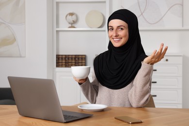 Photo of Muslim woman with cup of coffee using video chat on laptop at wooden table in room