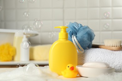 Photo of Baby cosmetic products, bath duck, brush and towel on white table against soap bubbles