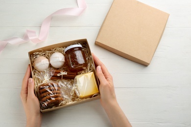 Woman holding box with stylish craft gift set at white wooden table, top view