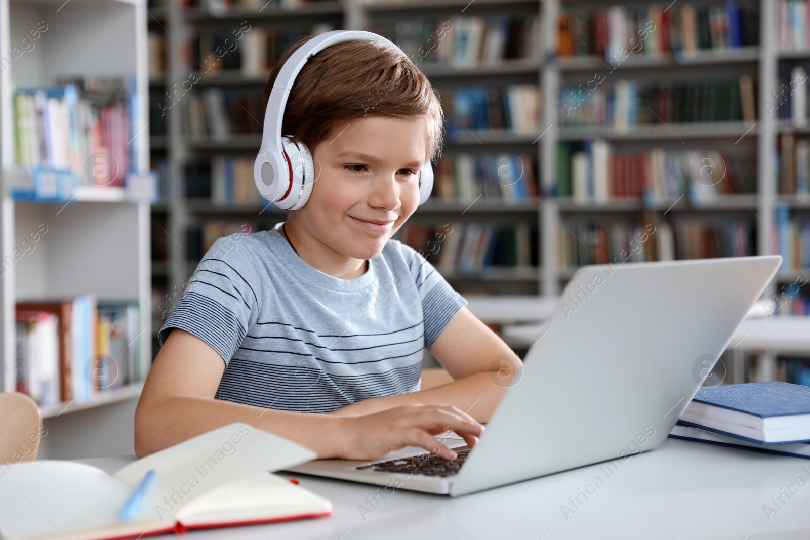 Photo of Little boy with headphones reading book using laptop in library