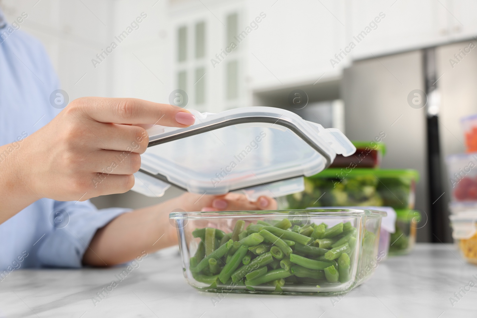 Photo of Woman sealing container with green beans at white marble table in kitchen, closeup. Food storage