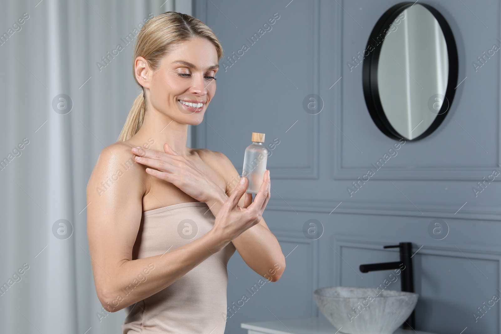 Photo of Happy woman with bottle of body oil in bathroom, space for text