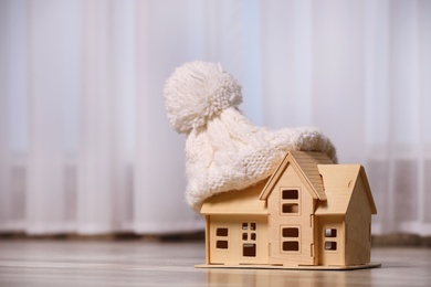 Photo of Wooden house model in hat on floor indoors, space for text. Heating efficiency