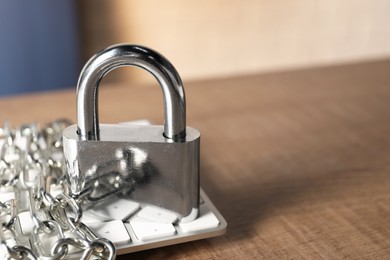 Photo of Cyber security. Keyboard with padlock and chain on wooden table, closeup. Space for text