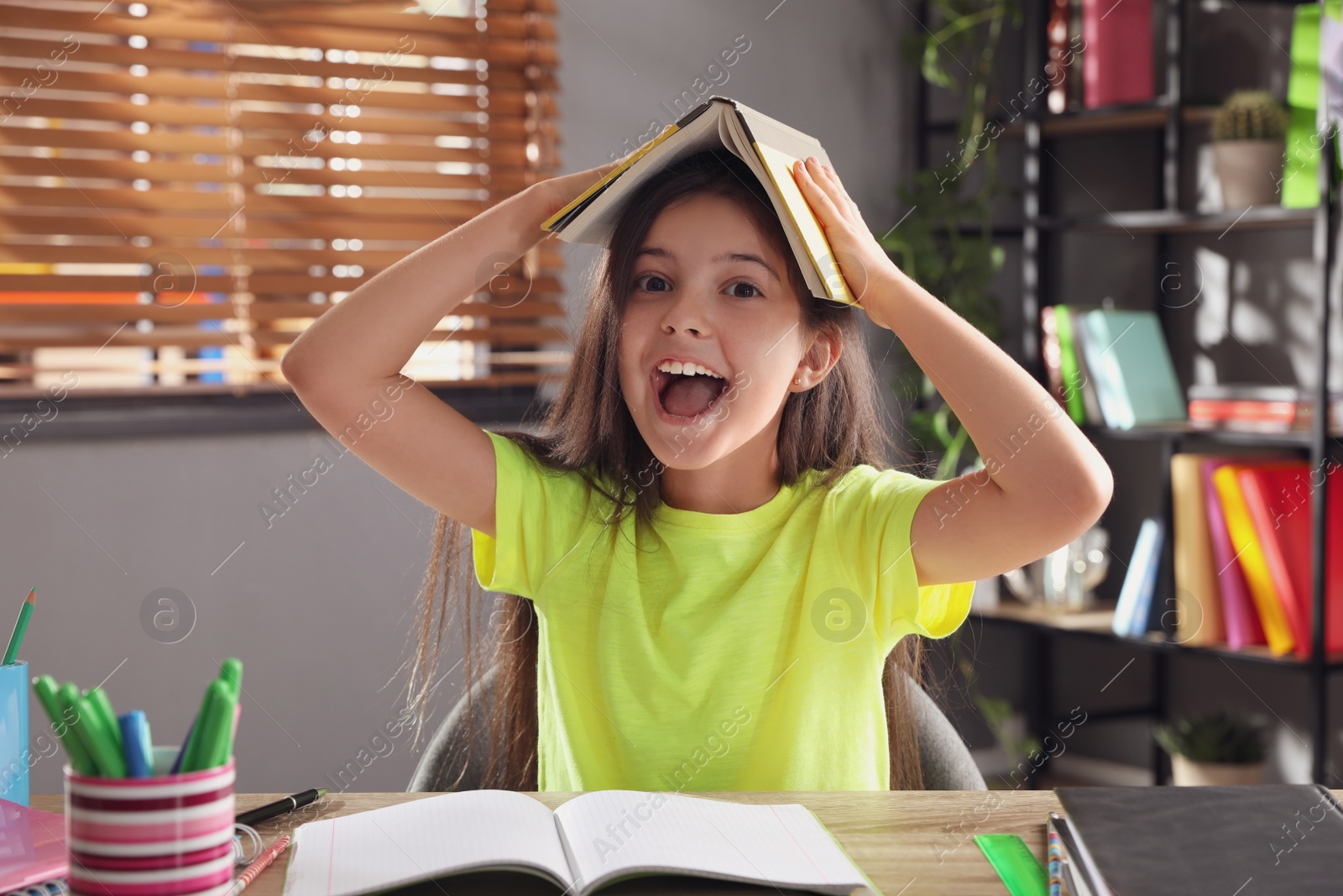 Photo of Emotional preteen girl playing with book at table. Doing homework