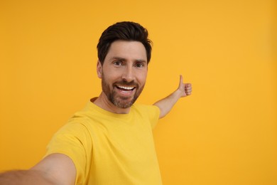 Photo of Smiling man taking selfie and showing thumbs up on yellow background, space for text