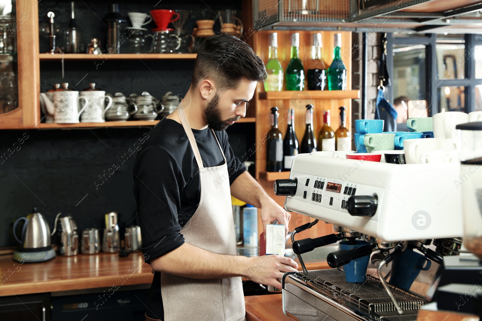 Photo of Barista pouring milk into metal jug near coffee machine at bar
