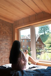 Young woman sitting on bed and enjoying view from window. Peaceful morning