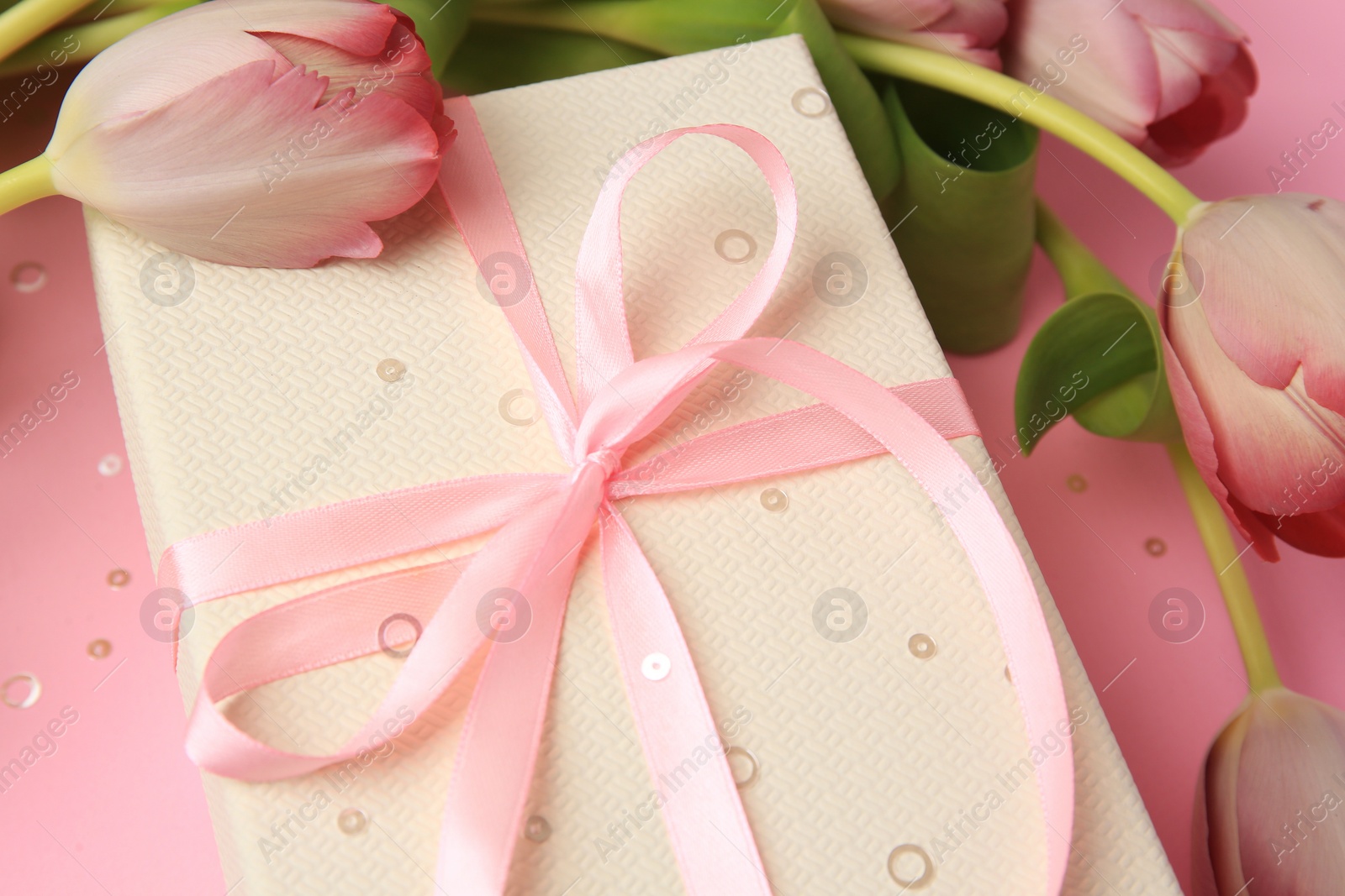 Photo of Beautiful gift box with bow, tulips and confetti on pink background, closeup