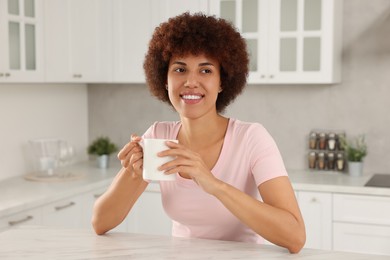 Photo of Happy young woman with cup of drink at table in kitchen