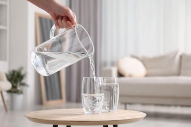 Woman pouring fresh water from jug into glass at wooden table indoors, closeup. Space for text