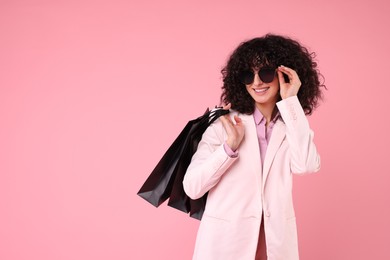 Happy young woman with shopping bags and stylish sunglasses on pink background