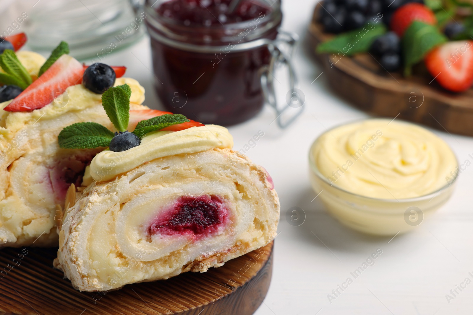 Photo of Pieces of tasty meringue roll with jam, cream, strawberry, blueberry and mint on white wooden table, closeup